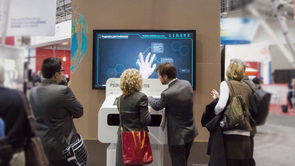 Photograph of a medical affairs conference booth exhibit. Two people are participating in an interactive activity about psoriatic arthritis using a kiosk. Three people are observing.