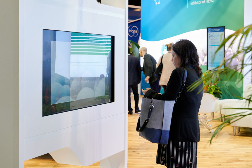 Photo a scientific animation displayed on a screen in a conference booth. A woman watches the video. © 2019 Seattle Genetics, Inc.