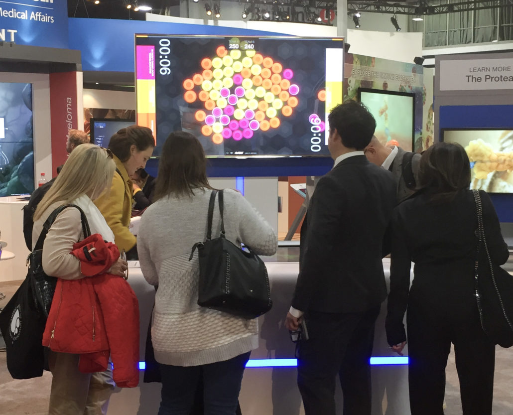 Photograph of multiplayer, interactive cancer pathophysiology touch-table game at a scientific conference booth. Two booth visitors (healthcare providers) are playing the game head to head, while four others look on in interest.