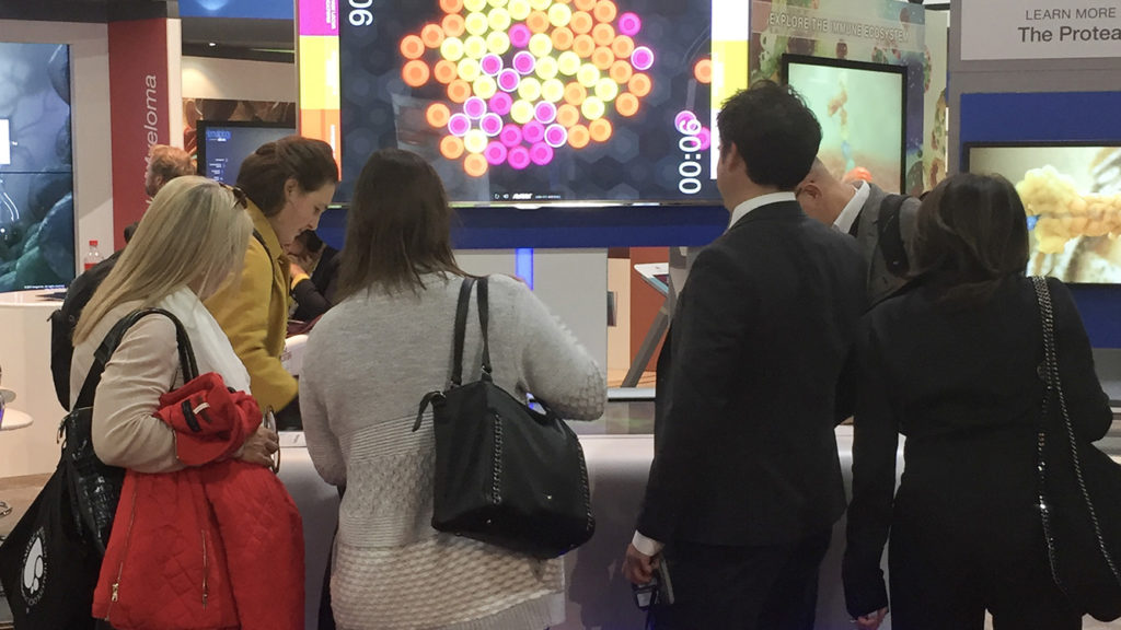 Amgen Oncology scientific affairs booth, ASH 2017, Atlanta, GA. Participants using the MM touch table with MM experience wall in the background. Photograph of multiplayer cancer pathophysiology touchtable game in a scientific conference booth. Two booth visitors (healthcare providers) are playing the game head to head, while four others look on in interest.