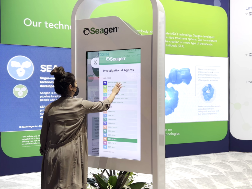 Photo of ASCO 2022 Medical Affairs booth. Woman uses interactive kiosk to learn about the drug development pipeline and clinical trials.