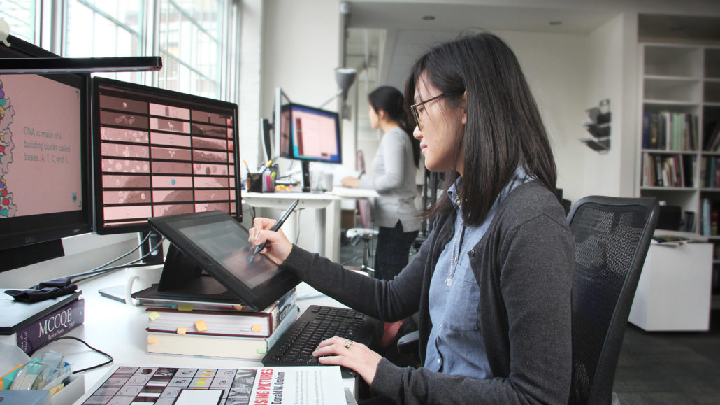 AXS Studio medical animators at work in a well-lit studio. The nearest animator is seated, with an array of screens including a storyboard of grey-pink tones and working with a stylus on a digital drawing tablet.
