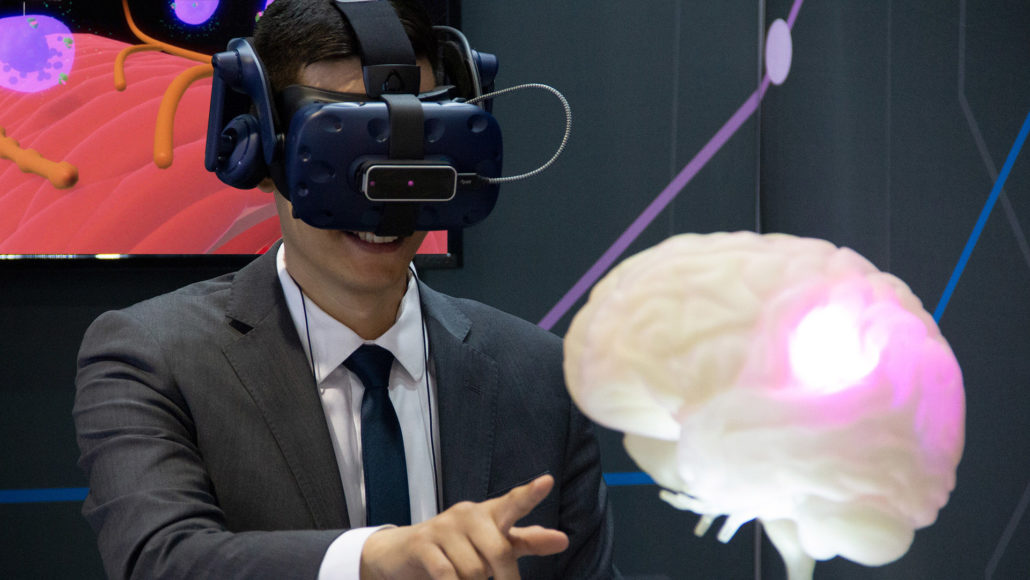 Photograph of VR user with Vive Pro head mounted display and Leap Motion controller. AXS Studio's 3D printed brain with LED lights, created for a Scientific Affairs booth at American Headache Society (AHS) 2019. Booth mural created by AXS Studio.