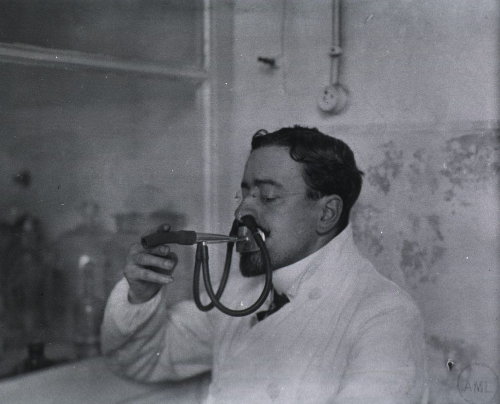 Black and white image of a male doctor with a goatee holding a device composed of rubber tubes and metal parts. Two tubes enter his nostrils and a plate covers his mouth.